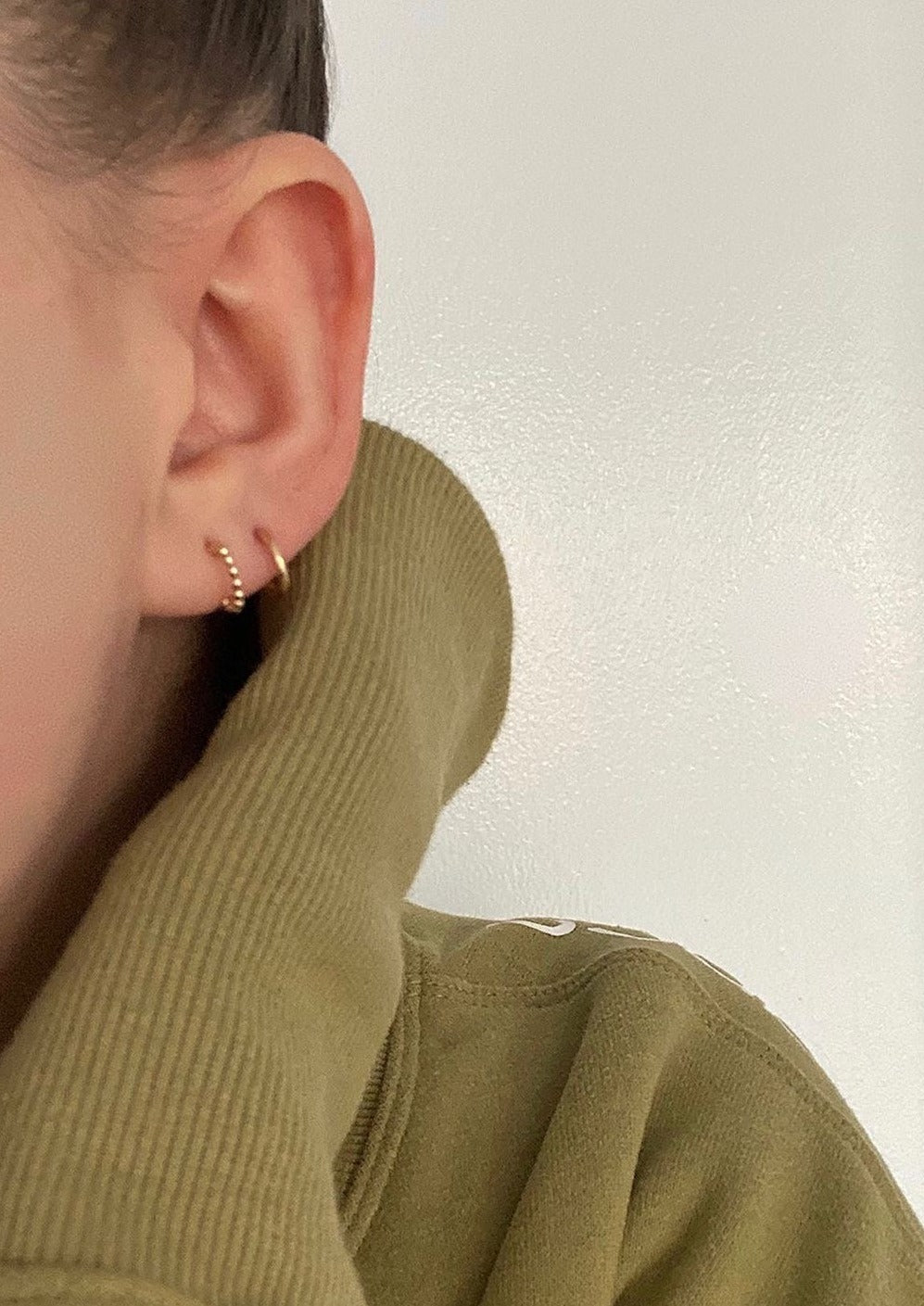 The model is wearing two pairs of the Ear Hugger Hoops in gold. One pair is smooth while the other is beaded. The model is wearing an olive green sweater.