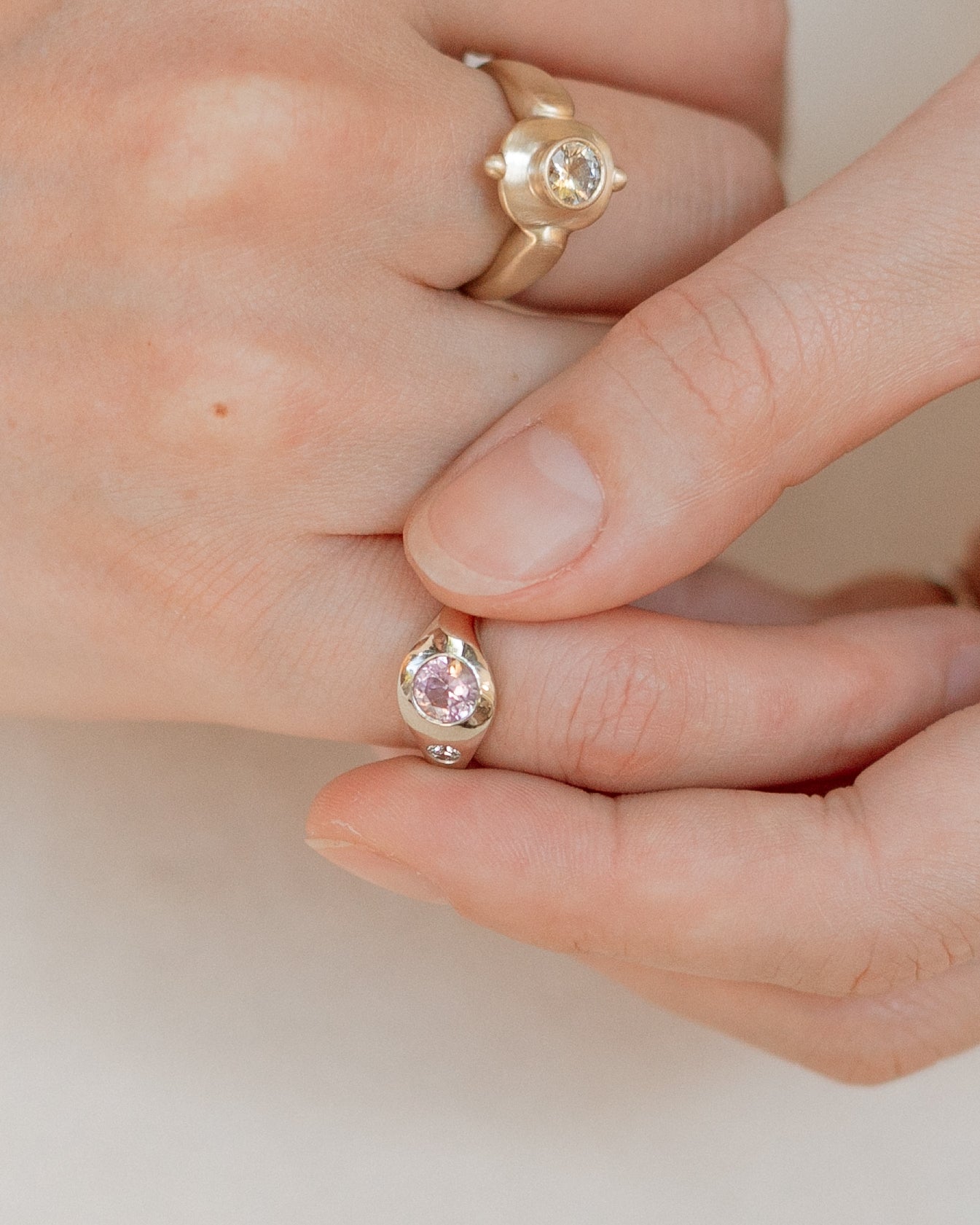 A close-up of a hand wearing two gold rings: a larger ring with a round clear stone on the back of the hand, and a smaller ring on the fingertip with a pink gemstone.