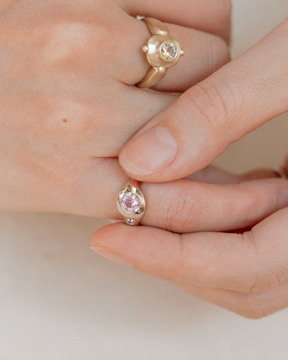 A close-up of a hand wearing two gold rings: a larger ring with a round clear stone on the back of the hand, and a smaller ring on the fingertip with a pink gemstone.