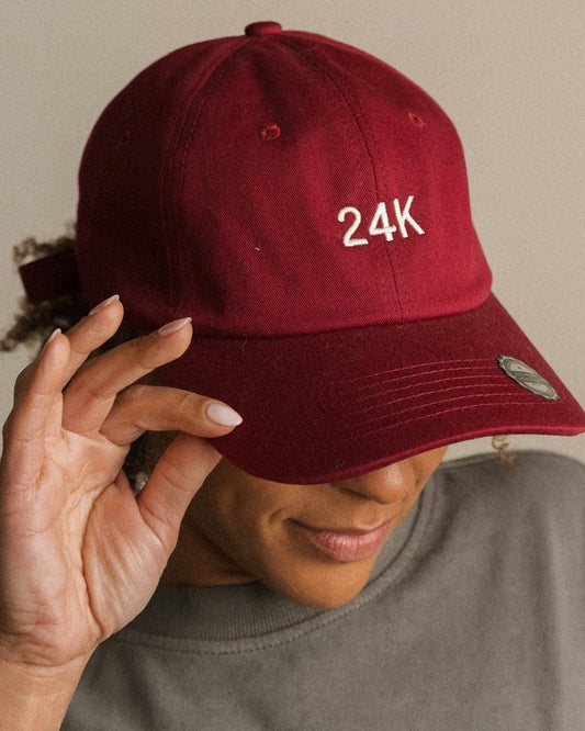 Person wearing a red cap with '24K' and a gray t-shirt with 'eye-YAH' text, sitting in front of a wall with 'Ayiah' sign.
